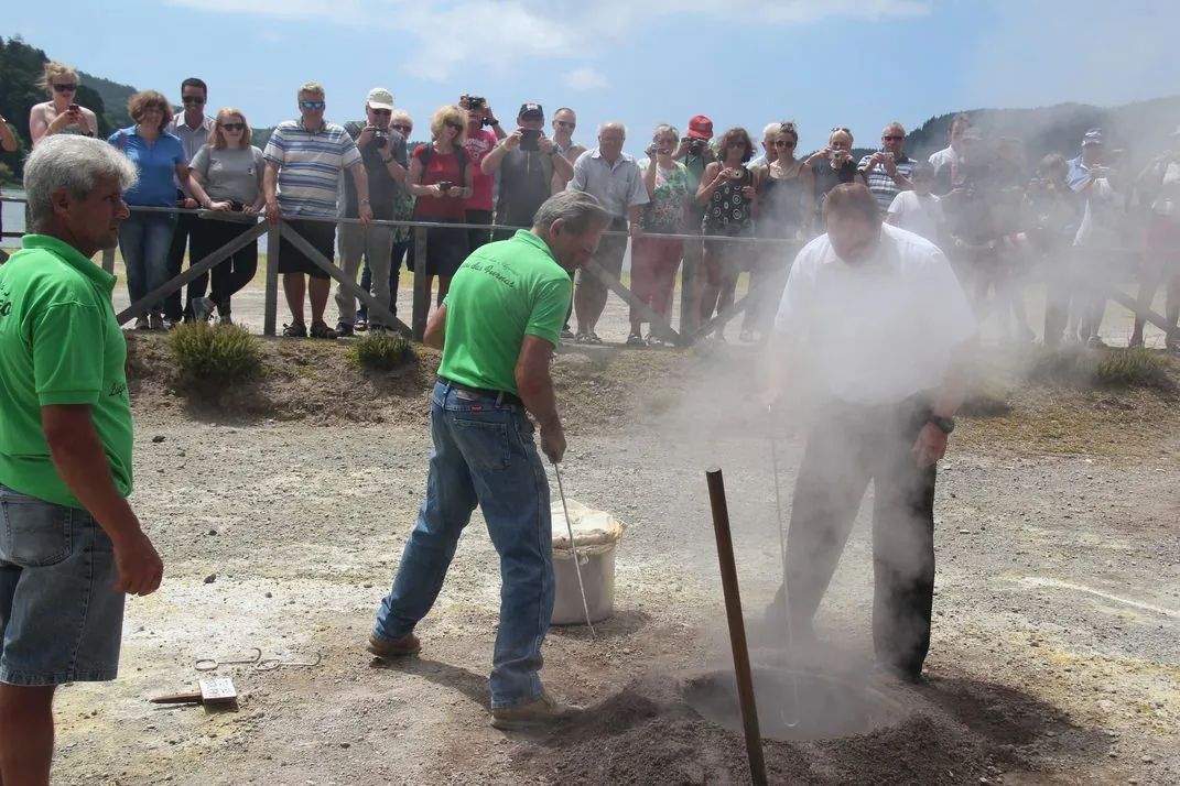 Cooks lifting a steaming pot from the volcanic ground at Furnas while visitors watch.