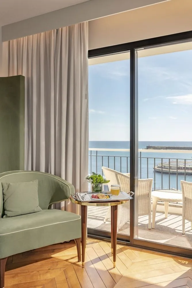 A seating area and bedroom beside a large harbour-facing window at Grand Hotel Açores Atlântico.